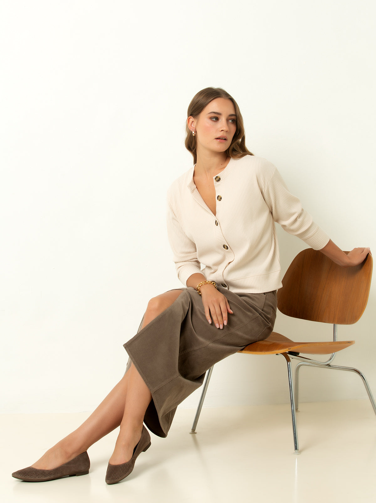 female model wearing a white colored ribbed cardigan sweater with buttons, aan d brown skirt and shoes sitting on a chair